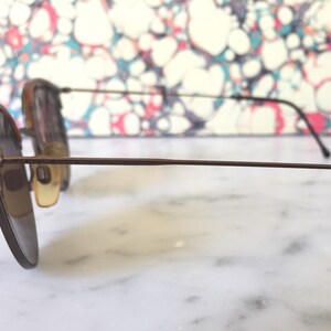 Puede incluir: Un par de gafas de sol con montura de metal marr&oacute;n y lentes marrones. Las gafas de sol est&aacute;n sobre una superficie blanca.