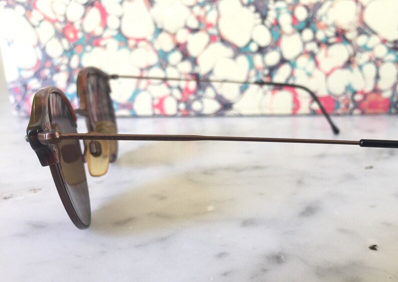 Puede incluir: Un par de gafas de sol con montura de metal marr&oacute;n y lentes marrones. Las gafas de sol est&aacute;n sobre una superficie blanca.