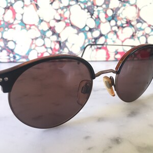 Puede incluir: Un par de gafas de sol redondas de color marr&oacute;n con montura de metal. Las gafas tienen un tinte marr&oacute;n y est&aacute;n sobre una superficie blanca.