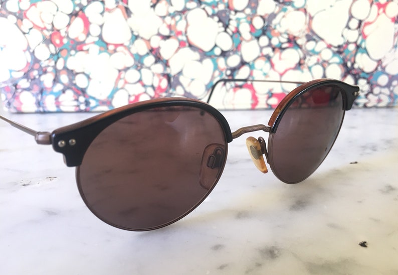 Puede incluir: Un par de gafas de sol redondas de color marr&oacute;n con montura de metal. Las gafas tienen un tinte marr&oacute;n y est&aacute;n sobre una superficie blanca.