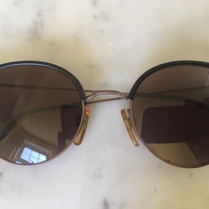 Puede incluir: Un par de gafas de sol redondas de color marr&oacute;n con monturas negras. Las monturas tienen un puente met&aacute;lico fino y las lentes est&aacute;n tintadas de marr&oacute;n.