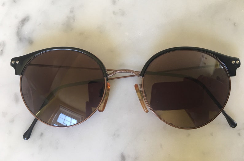 Puede incluir: Un par de gafas de sol redondas de color marr&oacute;n con monturas negras. Las monturas tienen un puente met&aacute;lico fino y las lentes est&aacute;n tintadas de marr&oacute;n.