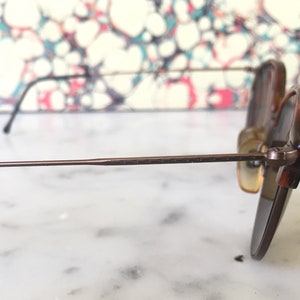 Puede incluir: Un par de gafas de sol con montura de metal marr&oacute;n y lentes marrones. Las gafas de sol est&aacute;n sobre una superficie de m&aacute;rmol blanco.