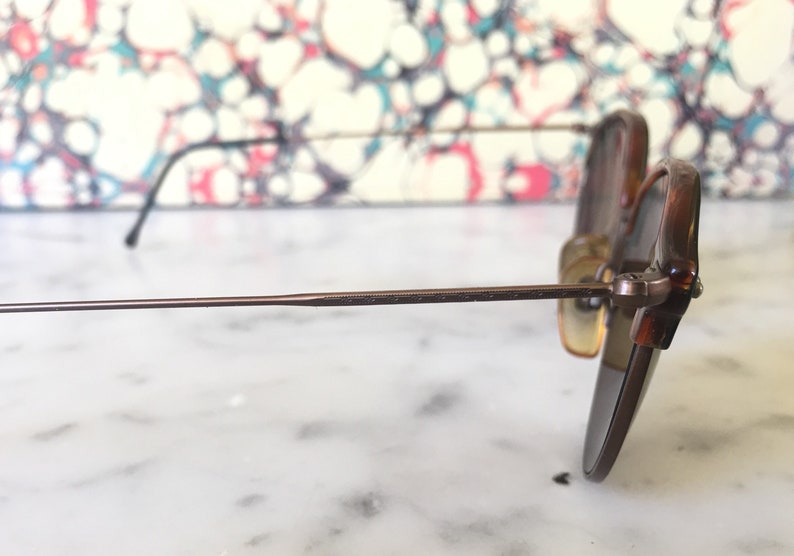 Puede incluir: Un par de gafas de sol con montura de metal marr&oacute;n y lentes marrones. Las gafas de sol est&aacute;n sobre una superficie de m&aacute;rmol blanco.