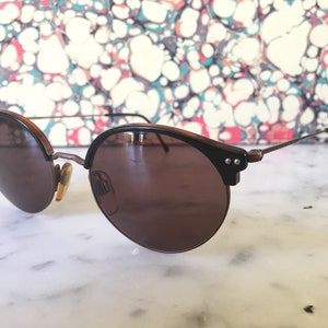 Puede incluir: Un par de gafas de sol redondas, de color marr&oacute;n, con un fino marco de metal dorado. Las gafas de sol tienen un borde de pl&aacute;stico marr&oacute;n oscuro alrededor de las lentes.