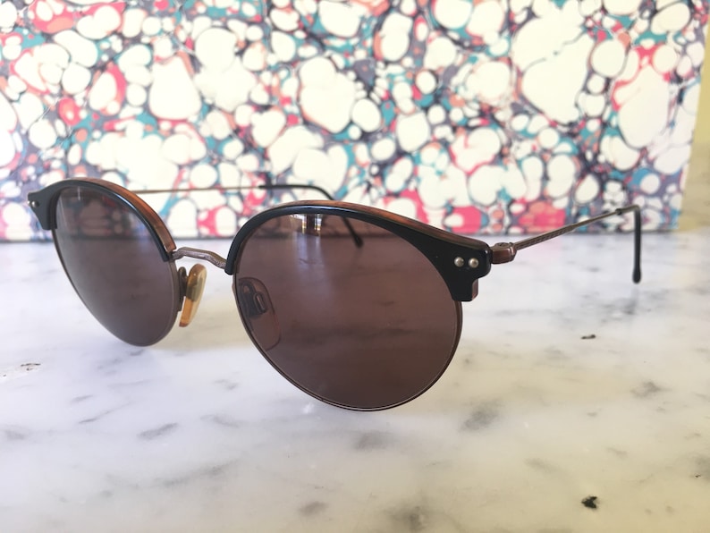 Puede incluir: Un par de gafas de sol redondas, de color marr&oacute;n, con un fino marco de metal dorado. Las gafas de sol tienen un borde de pl&aacute;stico marr&oacute;n oscuro alrededor de las lentes.