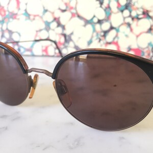 Puede incluir: Un par de gafas de sol redondas, de color marr&oacute;n, con monturas negras y detalles met&aacute;licos. Las gafas de sol tienen un estilo cl&aacute;sico y vintage.