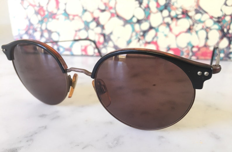 Puede incluir: Un par de gafas de sol redondas, de color marr&oacute;n, con monturas negras y detalles met&aacute;licos. Las gafas de sol tienen un estilo cl&aacute;sico y vintage.