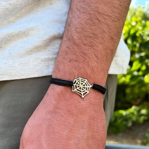 Peut inclure: Bracelet noir tressé avec un charm en forme de toile d'araignée de couleur or. Le bracelet est porté au poignet, mettant en valeur le design complexe de la toile. Un accessoire élégant.