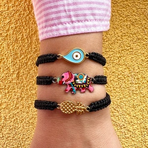May include: Three black string bracelets with charms. The top charm is a light blue teardrop with a white eye. The middle charm is a pink elephant with colorful patterns. The bottom charm is a gold pineapple.