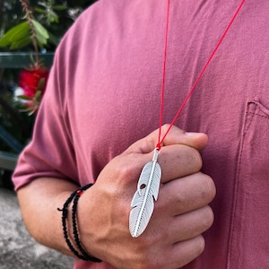 Könnte beinhalten: Halskette mit einem silbernen Federanhänger an einem roten Band. Der Anhänger hat geätzte Linien und ein kleines Loch. Ein schwarzes Perlenarmband ist ebenfalls zu sehen.