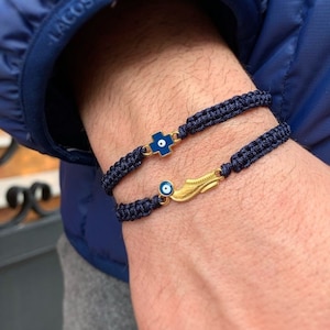Könnte beinhalten: Zwei blaue Kordelarmbänder mit goldenen Anhängern. Ein Anhänger ist ein blaues böses Auge mit einem goldenen Flügel, der andere ist ein blaues Kreuz.