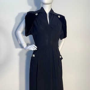 May include: A black vintage dress with a V-neckline and short sleeves. The dress has a fitted bodice and a full skirt. The shoulders have velvet accents and the dress is adorned with rhinestone buttons.