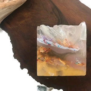May include: A clear, square soap bar with embedded gold flakes and colorful accents. The soap has a layered appearance with a pink, white, and yellow base.