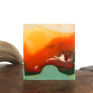 May include: A square-shaped bar of soap with a layered design. The top layer is a light yellow, the middle layer is orange, and the bottom layer is a teal green. The soap has a red and green swirl design in the middle.