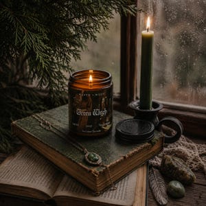 May include: A lit amber glass candle jar labelled Maven Moon Green Witch with 8 oz Coconut Soy Candle text, placed on a weathered green book next to a tall olive green taper candle in a black holder and a gold pendant necklace with a green stone.