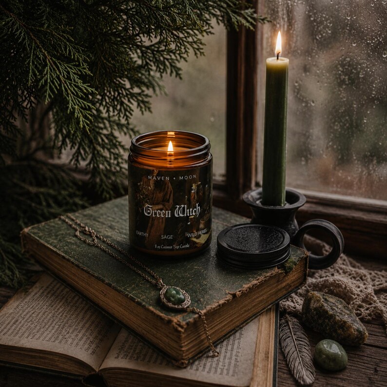 May include: A lit amber glass candle jar labelled Maven Moon Green Witch with 8 oz Coconut Soy Candle text, placed on a weathered green book next to a tall olive green taper candle in a black holder and a gold pendant necklace with a green stone.