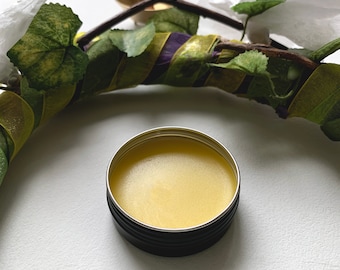 May include: A small round tin container with a black lid filled with a yellow balm or salve. The container is sitting on a white surface.