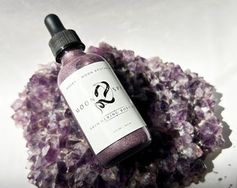 Puede incluir: Una pequeña botella de aceite corporal brillante con una etiqueta en blanco y negro que dice "Moon Spell Shimmering Body Oil" y "Maven Moon Apothecary". La botella está sentada sobre un lecho de cristales de amatista púrpura.