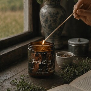 May include: An amber glass jar candle with a black label featuring the text Maven + Moon, Green Witch, Earthy, Sage, Wilderness, and 8 oz Coconut Soy Candle. The candle has a lit wick and is being lit by a wooden matchstick.