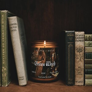 May include: A lit candle in a clear glass jar with a label reading Maven + Moon Green Witch Earthy Sage Wild Herbs 8 oz Coconut Soy Candle, positioned between two stacks of vintage books with titles like Gardening Encyclopedia and The Book of Garden Flowers.