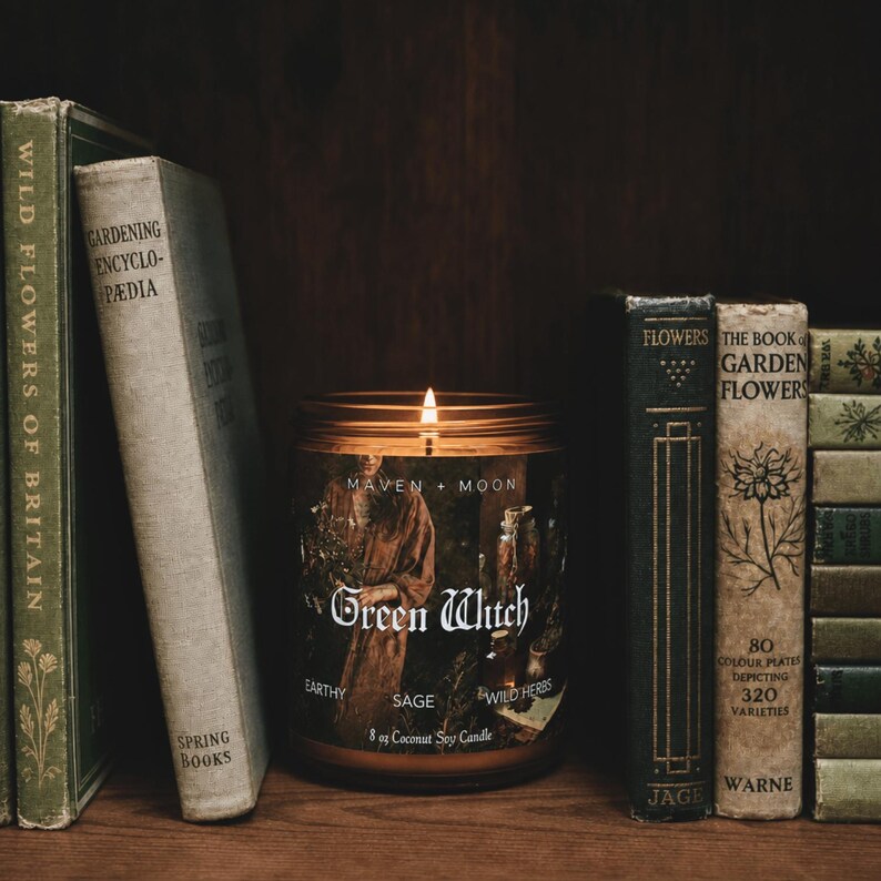May include: A lit candle in a clear glass jar with a label reading Maven + Moon Green Witch Earthy Sage Wild Herbs 8 oz Coconut Soy Candle, positioned between two stacks of vintage books with titles like Gardening Encyclopedia and The Book of Garden Flowers.