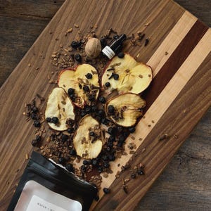 Könnte beinhalten: Ein hölzernes Schneidebrett zeigt getrocknete Apfelscheiben, eine ganze Muskatnuss, dunkle Beeren und kleine Gewürzstücke neben einer kleinen Tropfflasche mit der Aufschrift Summer Kitchen und einem schwarzen Beutel mit Maven + Muse.