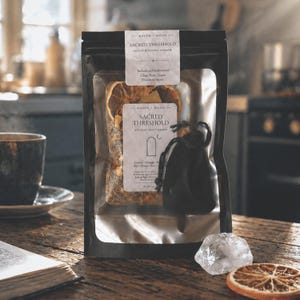 May include: A black package labeled "Sacred Threshold House Blessing Smimmer" containing dried herbs and citrus slices. A small black drawstring bag and a clear quartz crystal are next to the package. A cup of coffee and an open book are in the background.