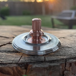 May include: A copper spinning top with a silver base. The top is on a wooden surface.
