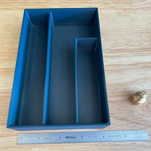 Puede incluir: Una bandeja rectangular azul marino con tres compartimentos y un pequeño peso de latón. Una regla está colocada junto a la bandeja. La bandeja mide unos 13 cm de largo.