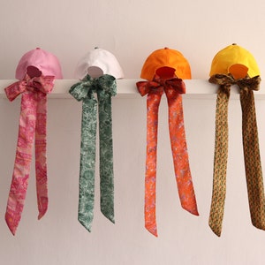 Puede incluir: Cuatro gorras de béisbol con diferentes tejidos de colores y lazos de cinta con patrones colgando de un estante blanco. Las gorras son rosa, blanca, naranja y amarilla.