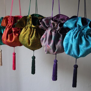 Könnte beinhalten: Sechs farbenfrohe Beutel mit Kordelzug aus glänzendem Stoff. Die Beutel sind in Türkis, Orange, Gold, Lila und Blau gehalten. Jeder Beutel hat eine passende Quaste und einen Kordelzugverschluss. Die Beutel hängen.