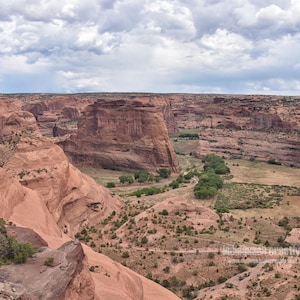 May include: A wide-angle view of a deep canyon featuring towering red sandstone cliffs and rock formations. The canyon floor is covered with patches of green trees and shrubs under a sky filled with heavy, grey, and white storm clouds.