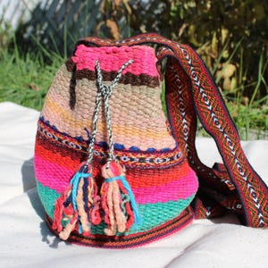 May include: A colorful woven drawstring bag with a long strap. The bag is made of a variety of colors, including pink, red, orange, yellow, green, and blue. The strap is woven with a geometric pattern in brown, red, and white.