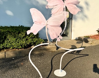 May include: Three decorative butterfly sculptures. Each butterfly has delicate pink wings and is attached to a white metal stand. The stands have a curved design and are placed on a paved surface. The butterflies appear to be made of a sheer fabric.