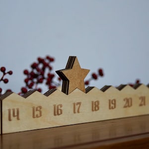 May include: A wooden advent calendar with numbers 14 to 21, a star on top, and red berries in the background.