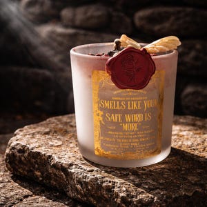 "Smells Like your safe word is "More"" 
Dark romance soy candle in matte white vessel with crimson wax bloom, golden wings, and gold foil label - Smells Like Your Safe Word Is More from Marked by Obsession collection