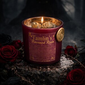 May include: A lit red candle in a glass jar with the text "Tamlin's Ravened Ruin" on a label. The candle has a gold rim and a gold seal. The candle is surrounded by red roses and sits on a stone.