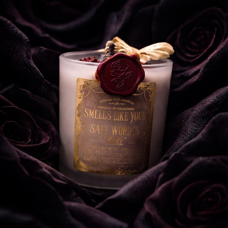 "Smells Like your safe word is "More"" 
Dark romance soy candle in matte white vessel with crimson wax bloom, golden wings, and gold foil label - Smells Like Your Safe Word Is More from Marked by Obsession collection