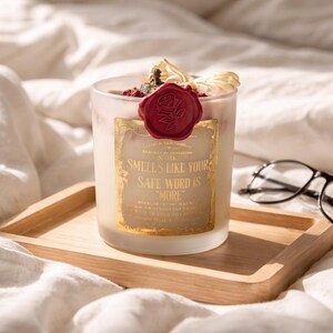 "Smells Like your safe word is "More"" 
Dark romance soy candle in matte white vessel with crimson wax bloom, golden wings, and gold foil label - Smells Like Your Safe Word Is More from Marked by Obsession collection