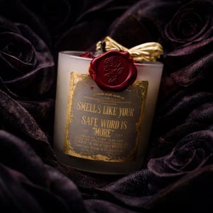 "Smells Like your safe word is "More"" 
Dark romance soy candle in matte white vessel with crimson wax bloom, golden wings, and gold foil label - Smells Like Your Safe Word Is More from Marked by Obsession collection
