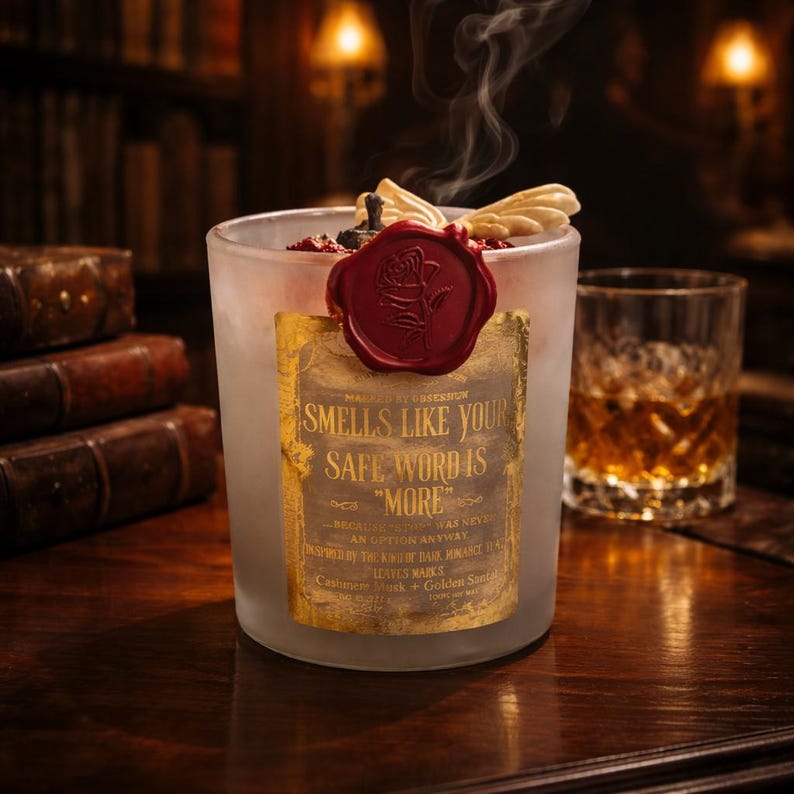 "Smells Like your safe word is "More"" 
Dark romance soy candle in matte white vessel with crimson wax bloom, golden wings, and gold foil label - Smells Like Your Safe Word Is More from Marked by Obsession collection