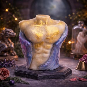 May include: A sculpted candle in the shape of a male torso. The candle is primarily cream-colored with gold accents on the chest and shoulders. The torso is draped with blue and purple wings, and sits on a dark gray square base.