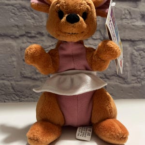 May include: A plush kangaroo toy with brown fur, pink inner ears, and a pink torso section. The kangaroo wears a white apron around its waist and has a black nose and eyes. A product tag is attached to the side, and a care label is visible at the base.