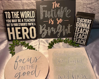 May include: A collection of decorative signs with inspirational quotes. The signs are in various shapes and sizes, with text such as "The Future is so Bright" and "Focus on the Good." Colors include black, white, and pink.