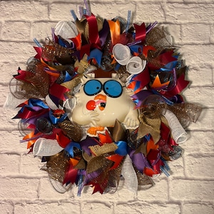 May include: A fall-themed wreath with a white ceramic owl figurine in the center. The wreath is decorated with brown, red, orange, blue, and purple ribbons, as well as brown burlap and white mesh.