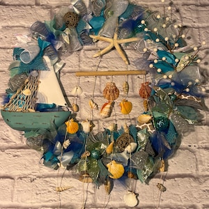 May include: A blue and white nautical themed wreath with a wooden sailboat, seashells, starfish, and white beads. The wreath is decorated with blue, white, and silver mesh and ribbons.