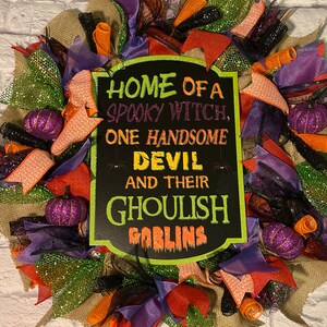 May include: A Halloween wreath featuring a black chalkboard sign that reads "Home of a spooky witch, one handsome devil and their ghoulish goblins". The wreath is decorated with orange, purple, green and black ribbons and mesh.