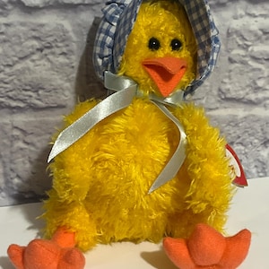 May include: A plush yellow chick wearing a blue and white gingham bonnet and a silver ribbon. The chick has an orange beak and feet. The stuffed animal is sitting on a white surface in front of a gray brick wall.