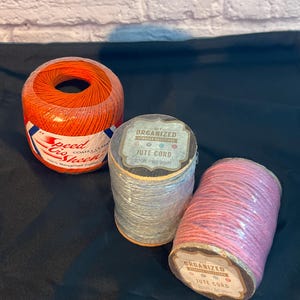 May include: Three spools of thread and cord. An orange spool of Speed Cro-Sheen thread is on the left. In the center is a spool of gray jute cord, and on the right is a pink spool of jute cord. Each spool is labeled.